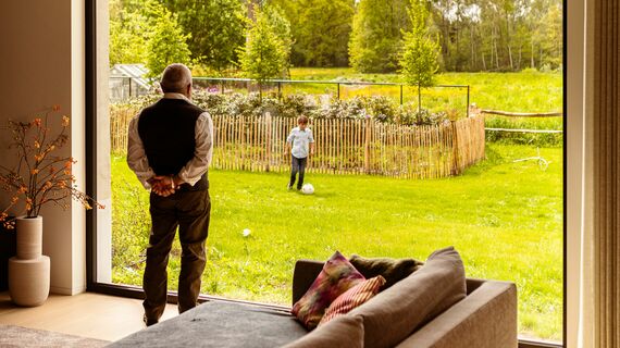 Elder person watching his grandson play in the garden from behind window wall.